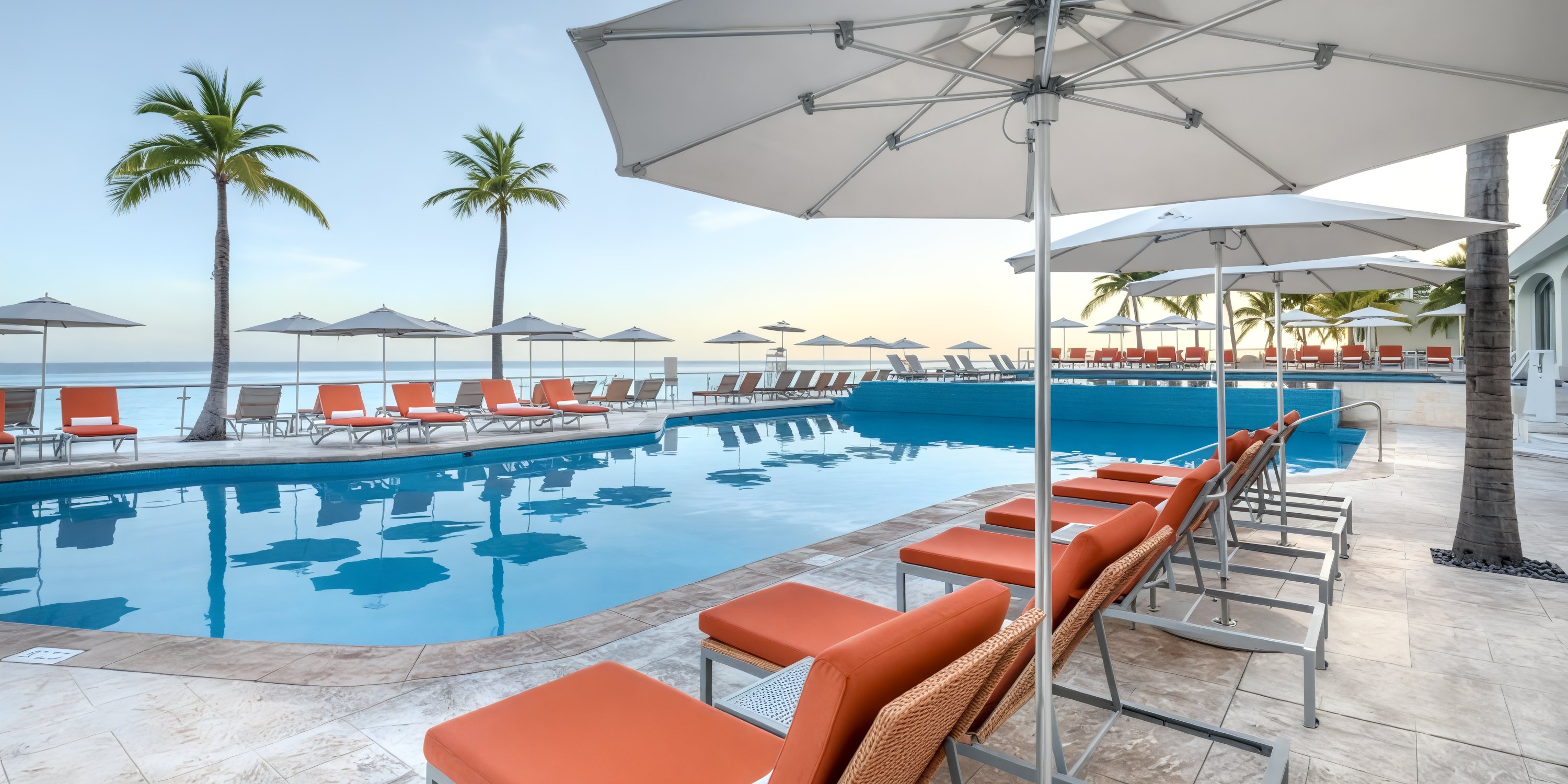 Cozumel Palace: An ideal destination wedding location featuring a poolside scene with vibrant orange loungers, elegant umbrellas, and ocean-view palm trees.