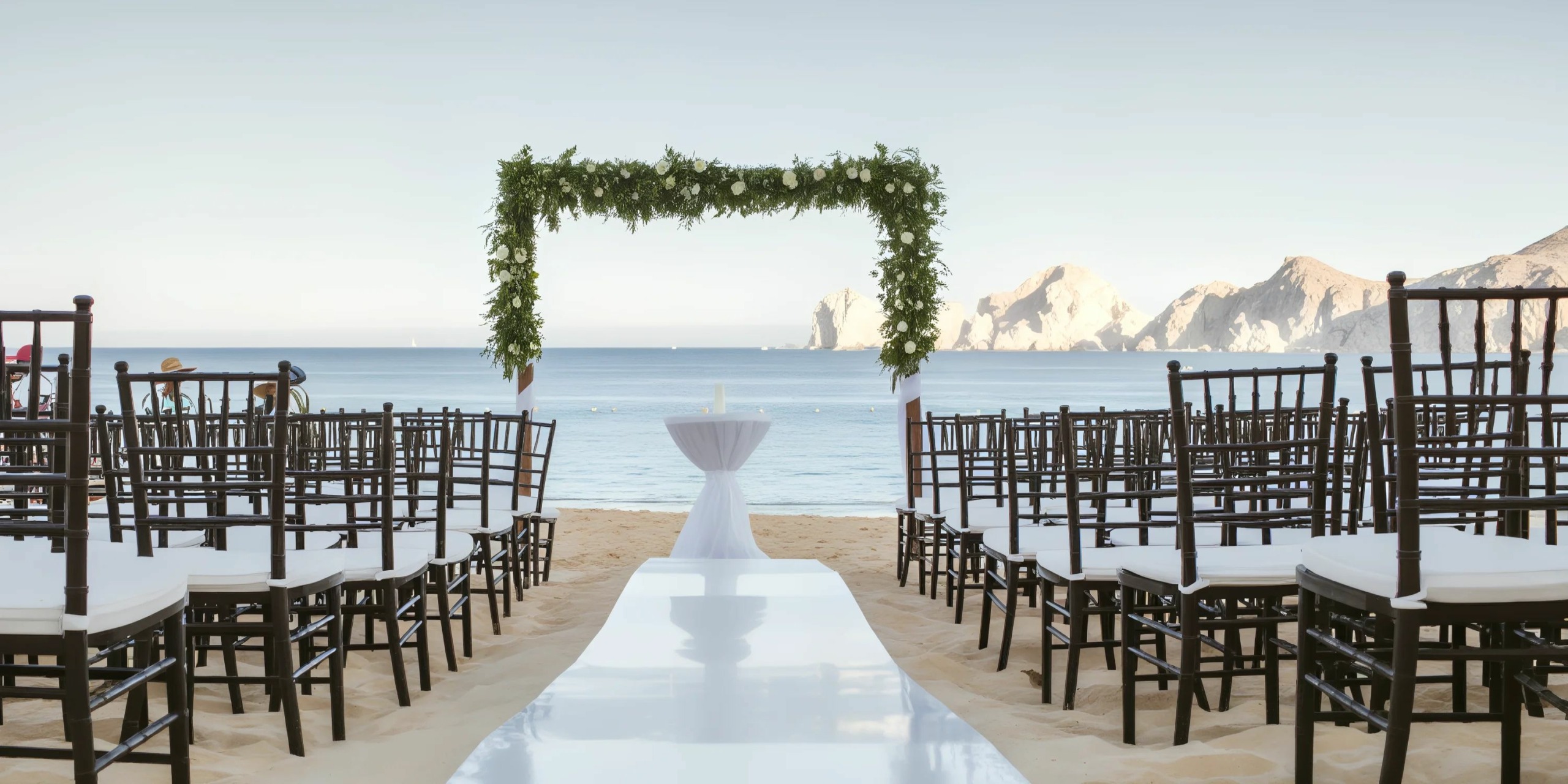 Outdoor beach wedding setup with a white aisle, black chairs on either side, and a green floral arch overlooking the ocean and distant rocky cliffs.