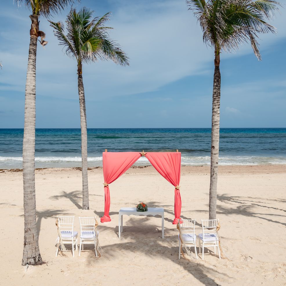 A destination wedding altar with pink drapes stands on a sandy beach framed by two palm trees.