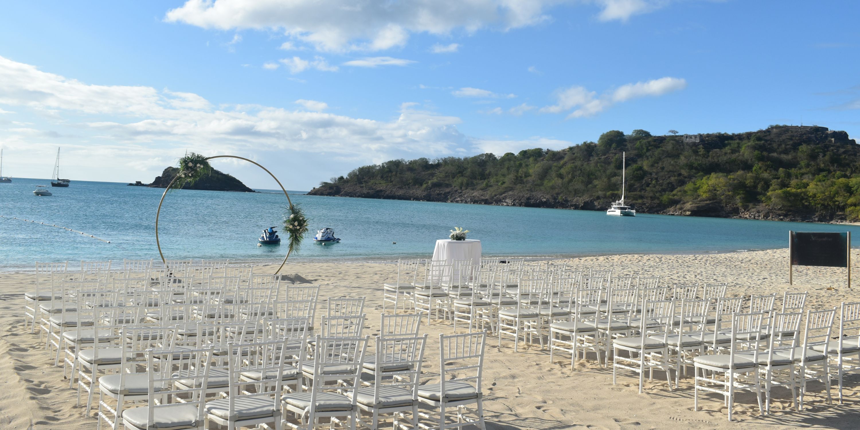 Rows of white chairs set up for a destination wedding face an arch on the beach at Royalton Antigua.