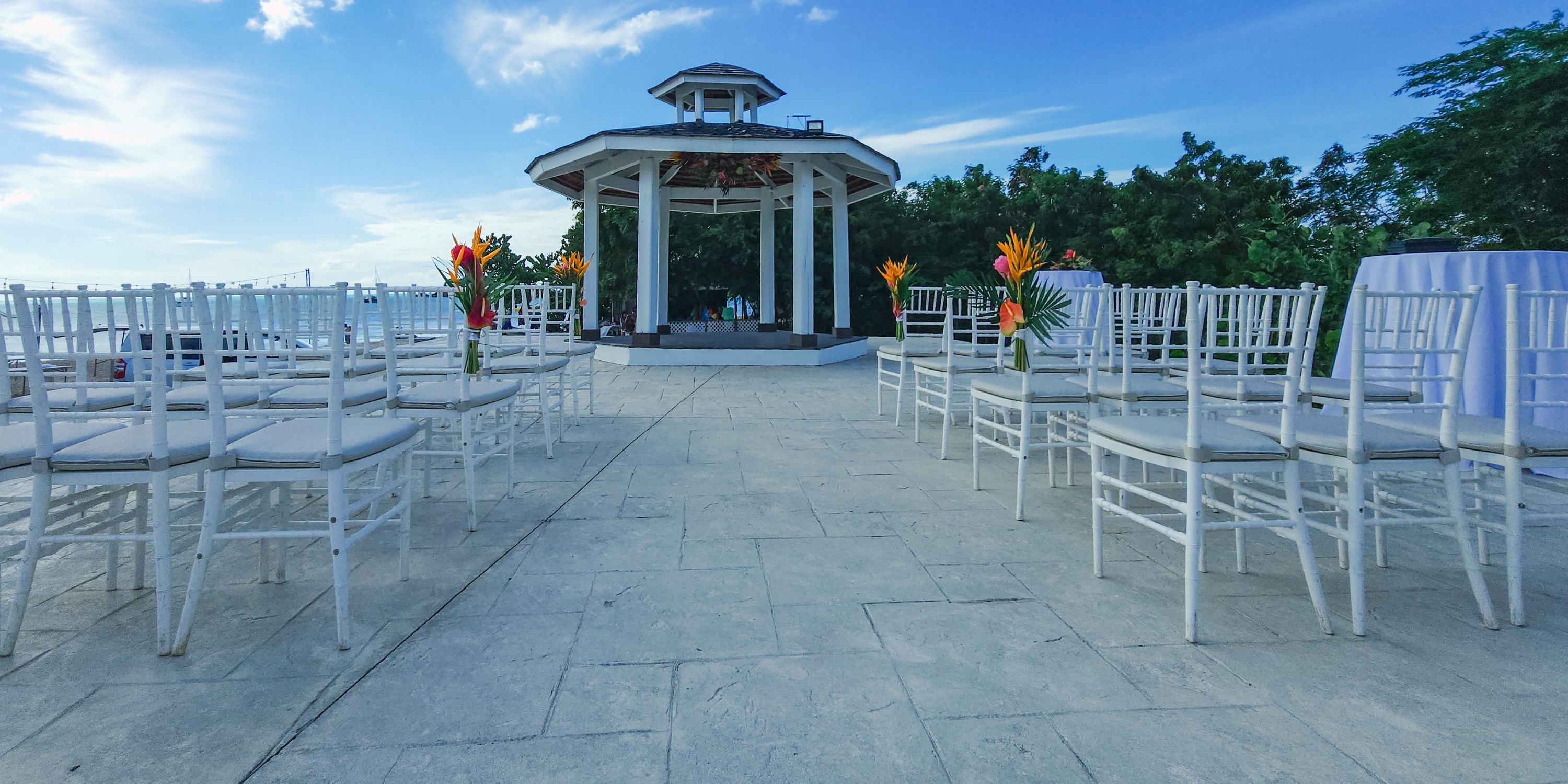 White chairs arranged for a destination wedding face a gazebo, tropical flowers adorning the aisle.