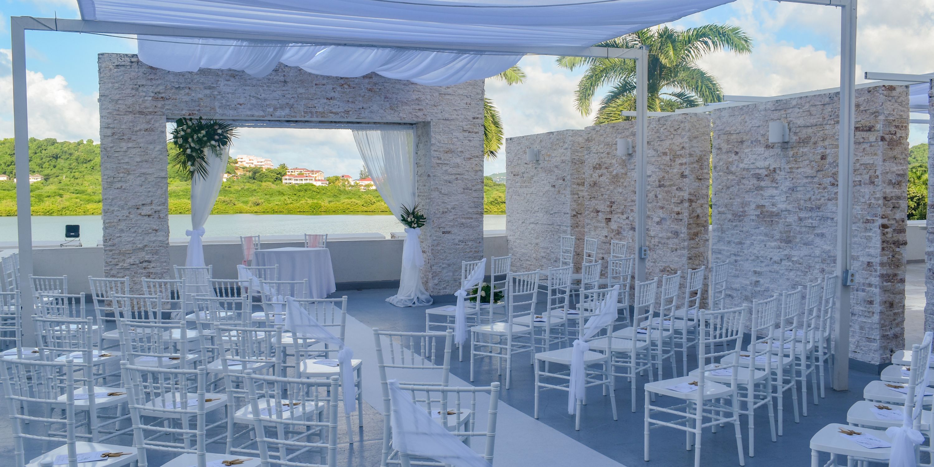 Destination wedding at Royalton Antigua featuring white chairs, stone arches, and fabric draping by water.