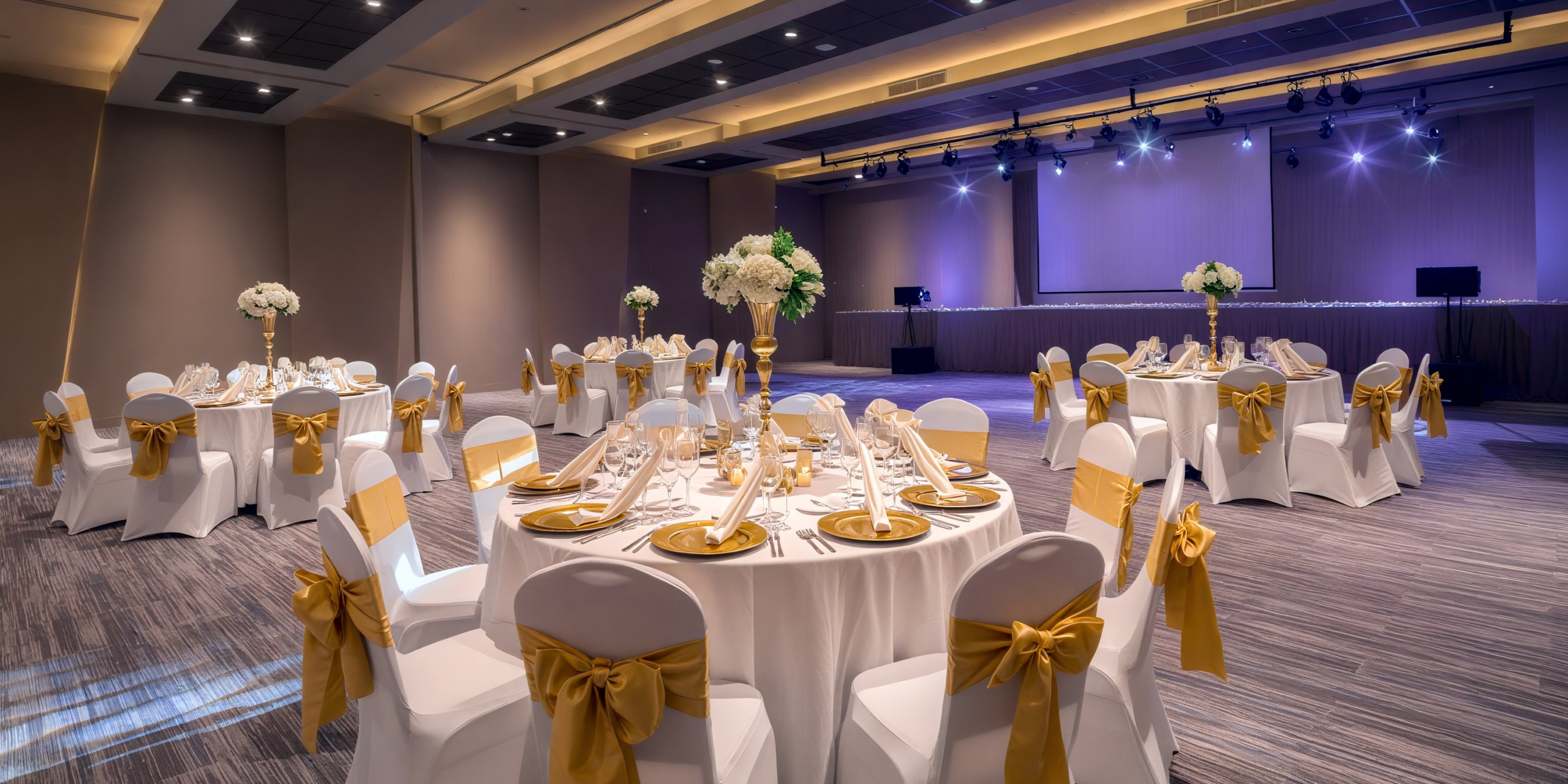 Royalton Antigua Hall is the perfect setting for a destination wedding, featuring round tables and elegant white chairs adorned with gold sashes. The stage, illuminated with blue lighting, creates a magical ambiance for your special day.