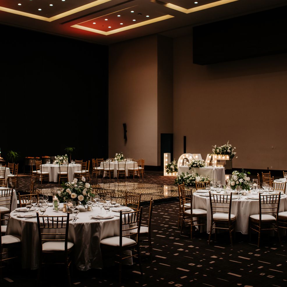 Spacious banquet hall prepared for a destination wedding, featuring round tables with white linens and floral centerpieces.