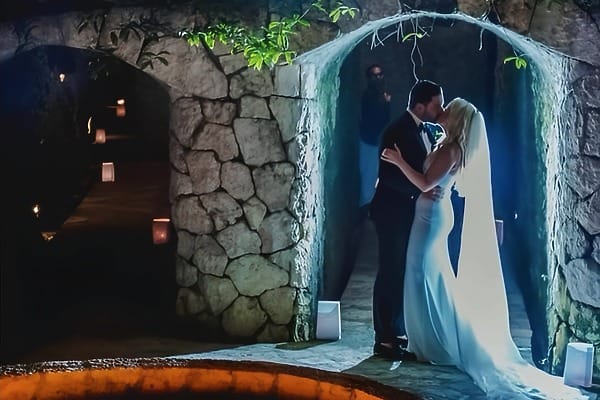 The couple shares a wedding kiss beneath a stone arch at Hotel Xcaret, under dramatic lighting.
