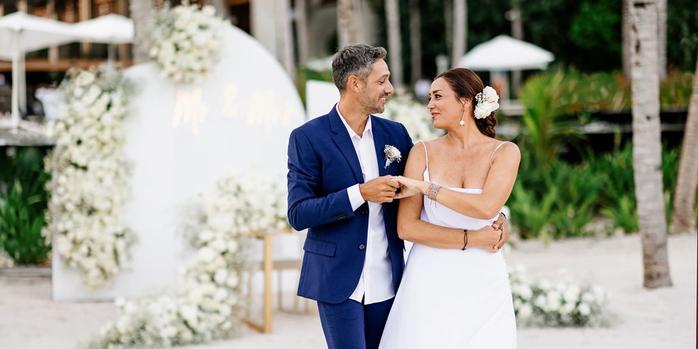 A couple in wedding attire beams at each other amidst a picturesque outdoor setting, enhanced by floral decorations, at the Hilton Tulum Riviera Maya All-Inclusive Resort.