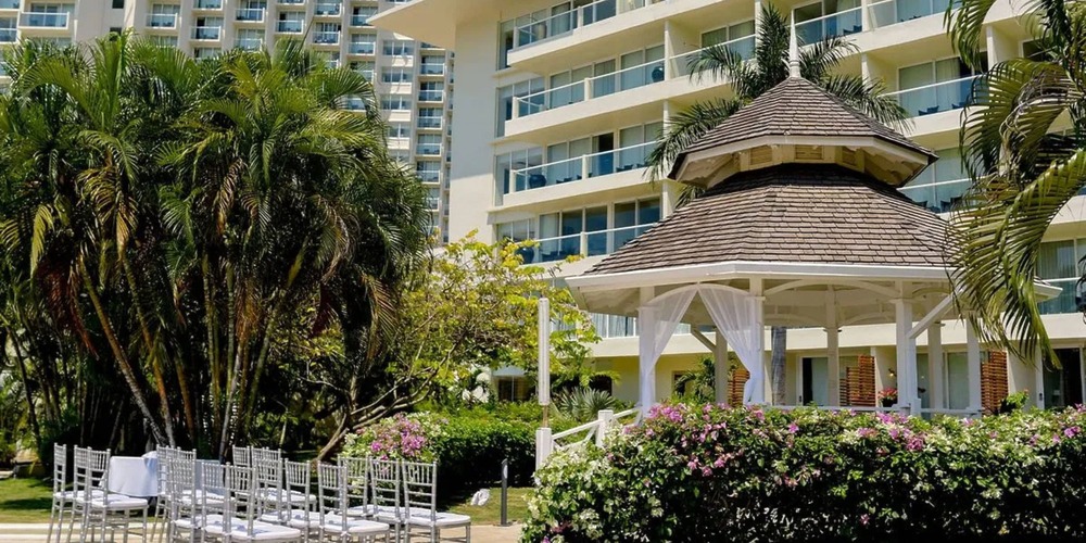 Outdoor wedding setup with white chairs facing a gazebo, surrounded by lush greenery, in front of the Moon Palace Jamaica building adorned with multiple balconies.
