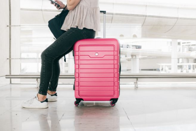 Woman in black jeans reads her phone while sitting on a pink roller luggage