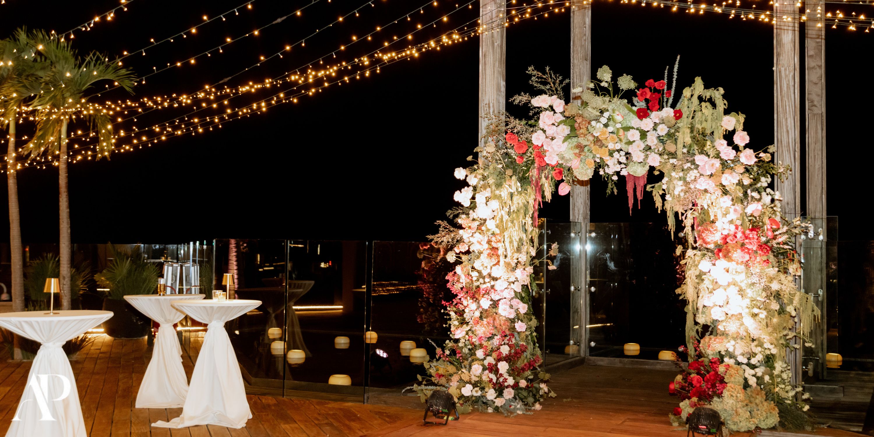 Wedding arch adorned with flowers, string lights, and cocktail tables at night for a destination wedding.