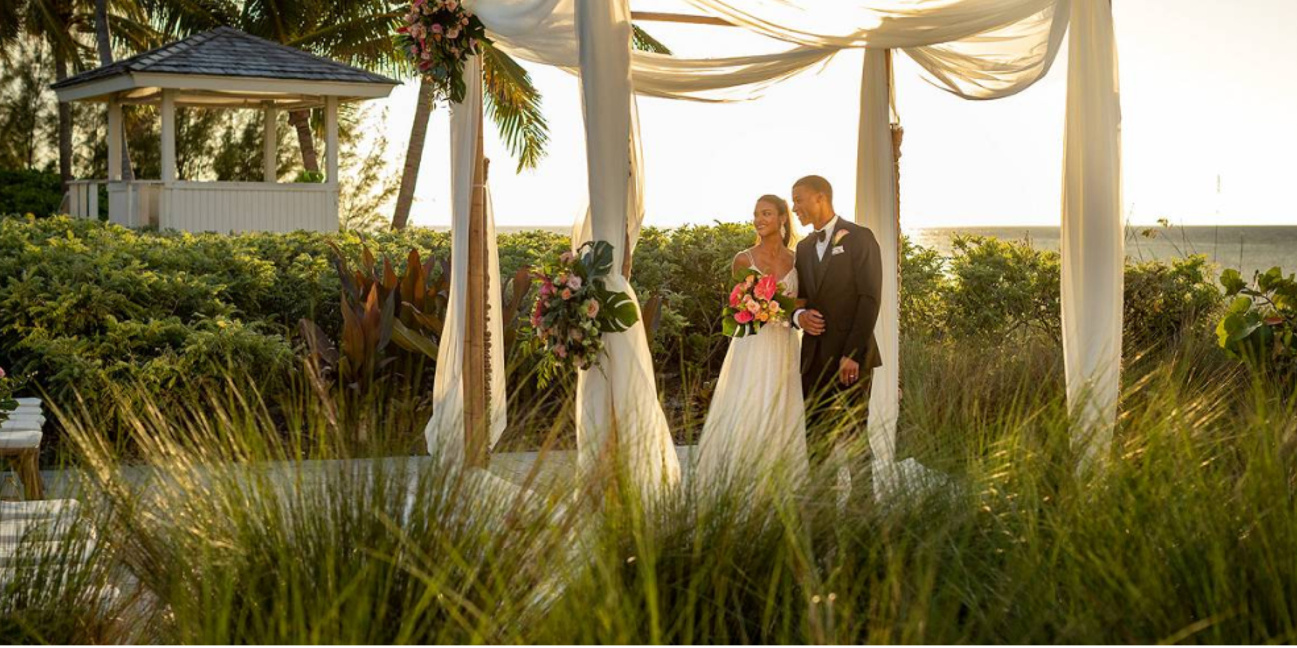 Newly married gorgeous couple stand hand in hand under a wedding venue, surrounded by a tropical garden.