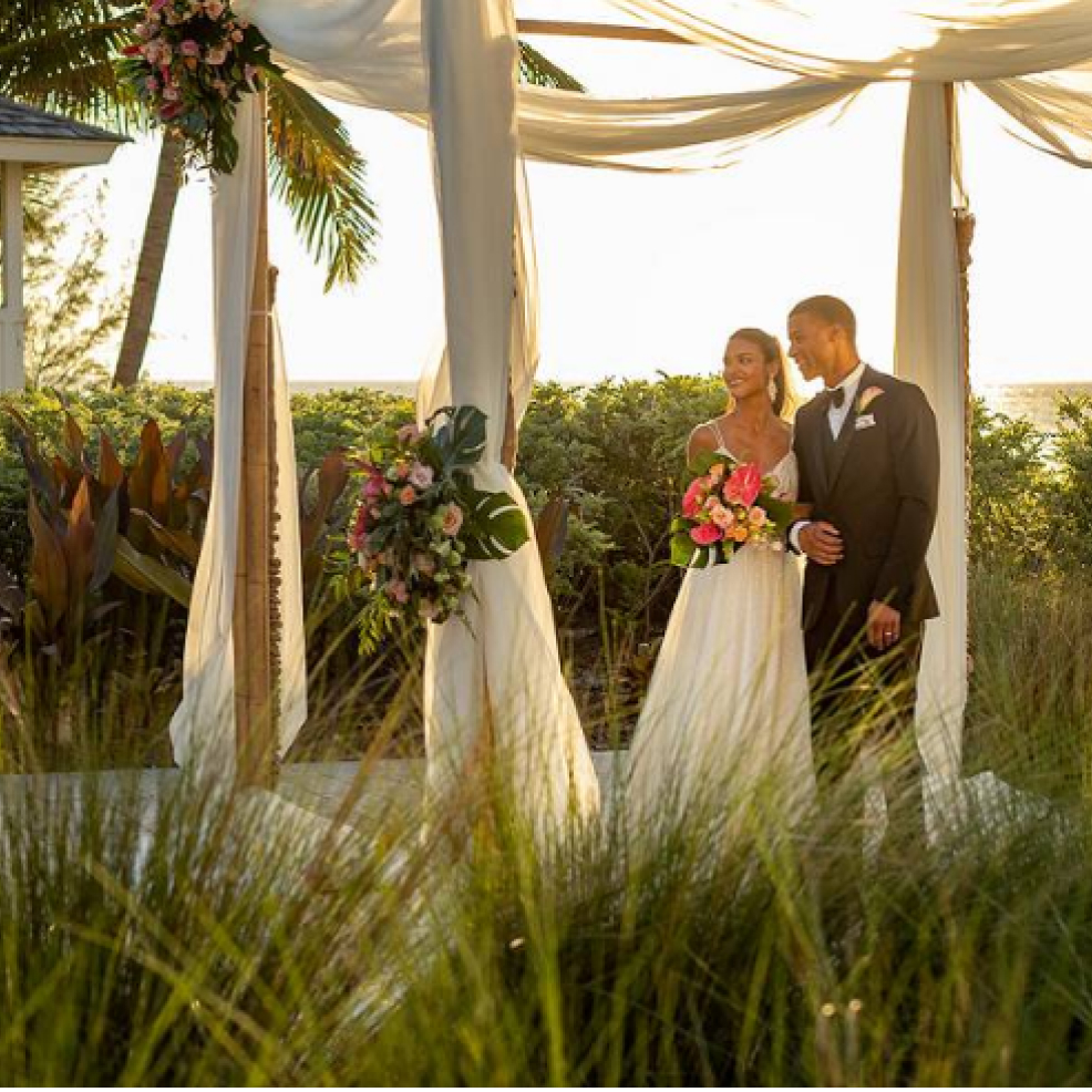 Newly married gorgeous couple stand hand in hand under a wedding venue, surrounded by a tropical garden.