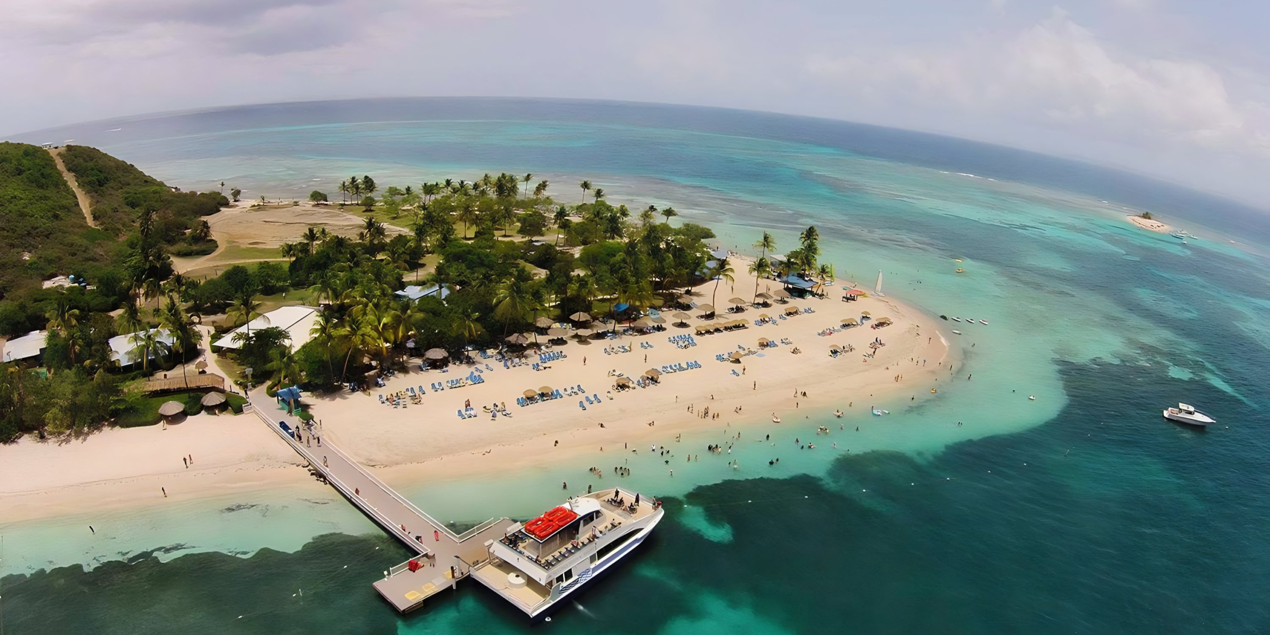 Aerial view of a tropical island, a popular destination for weddings, with a sandy beach, boats near shore, and lush greenery surrounded by turquoise water.