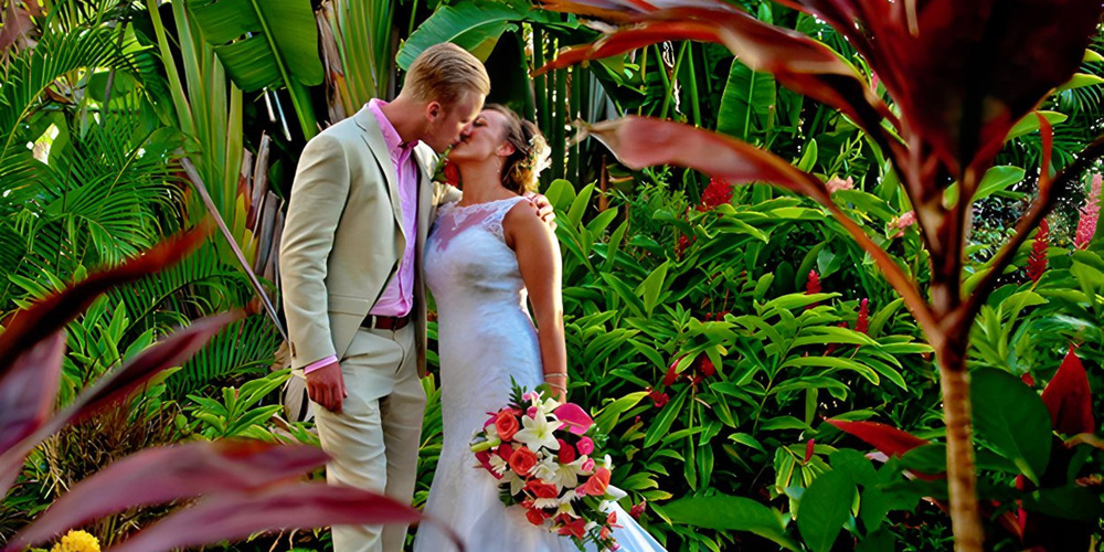 A couple in wedding attire share a kiss in a lush, tropical garden surrounded by vibrant green foliage and colorful flowers. The bride holds a bouquet of pink and white flowers, epitomizing the romance promised by Barbados Wedding Packages.