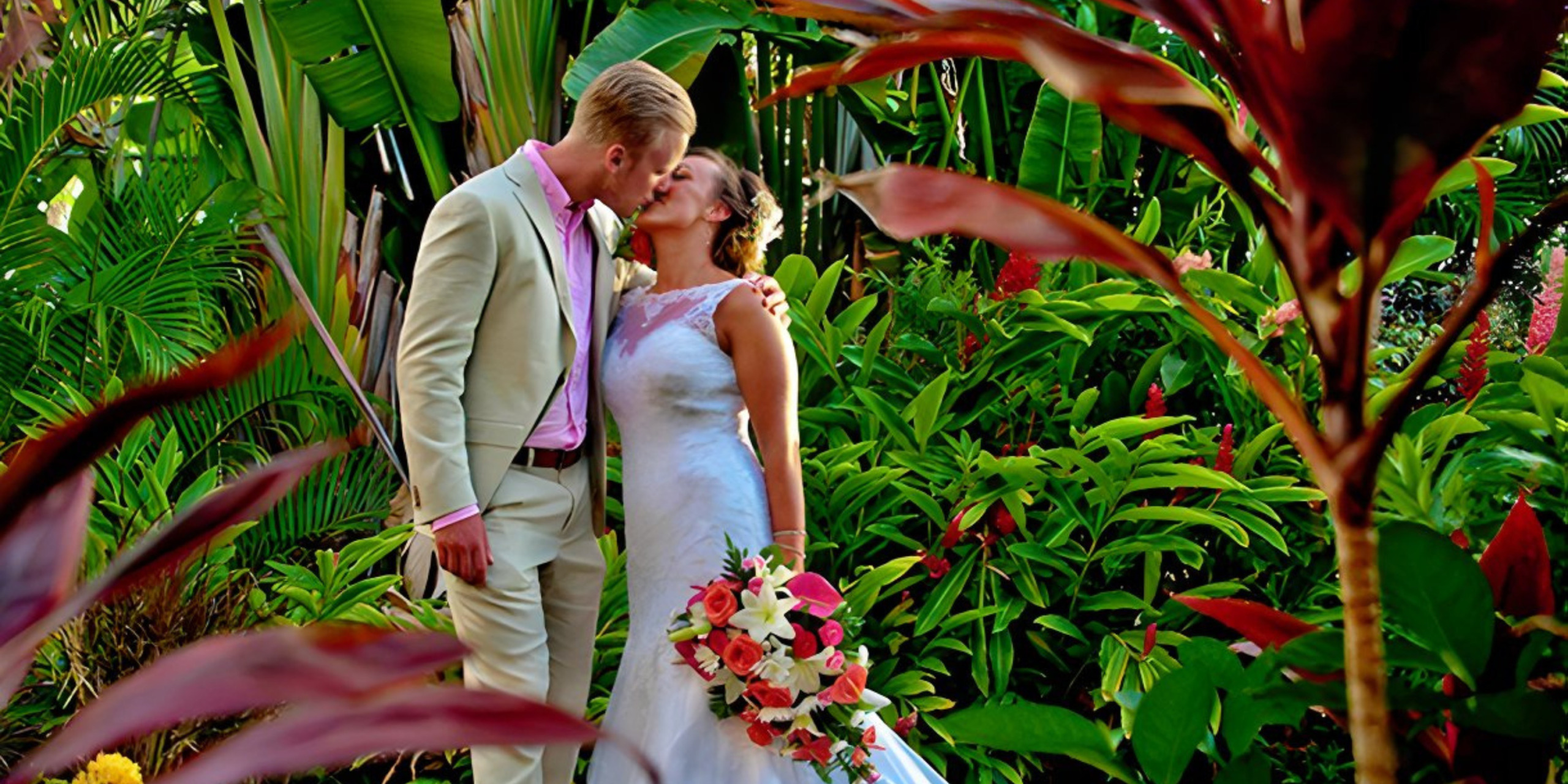 A couple in wedding attire share a kiss in a lush, tropical garden surrounded by vibrant green foliage and colorful flowers. The bride holds a bouquet of pink and white flowers, epitomizing the romance promised by Barbados Wedding Packages.