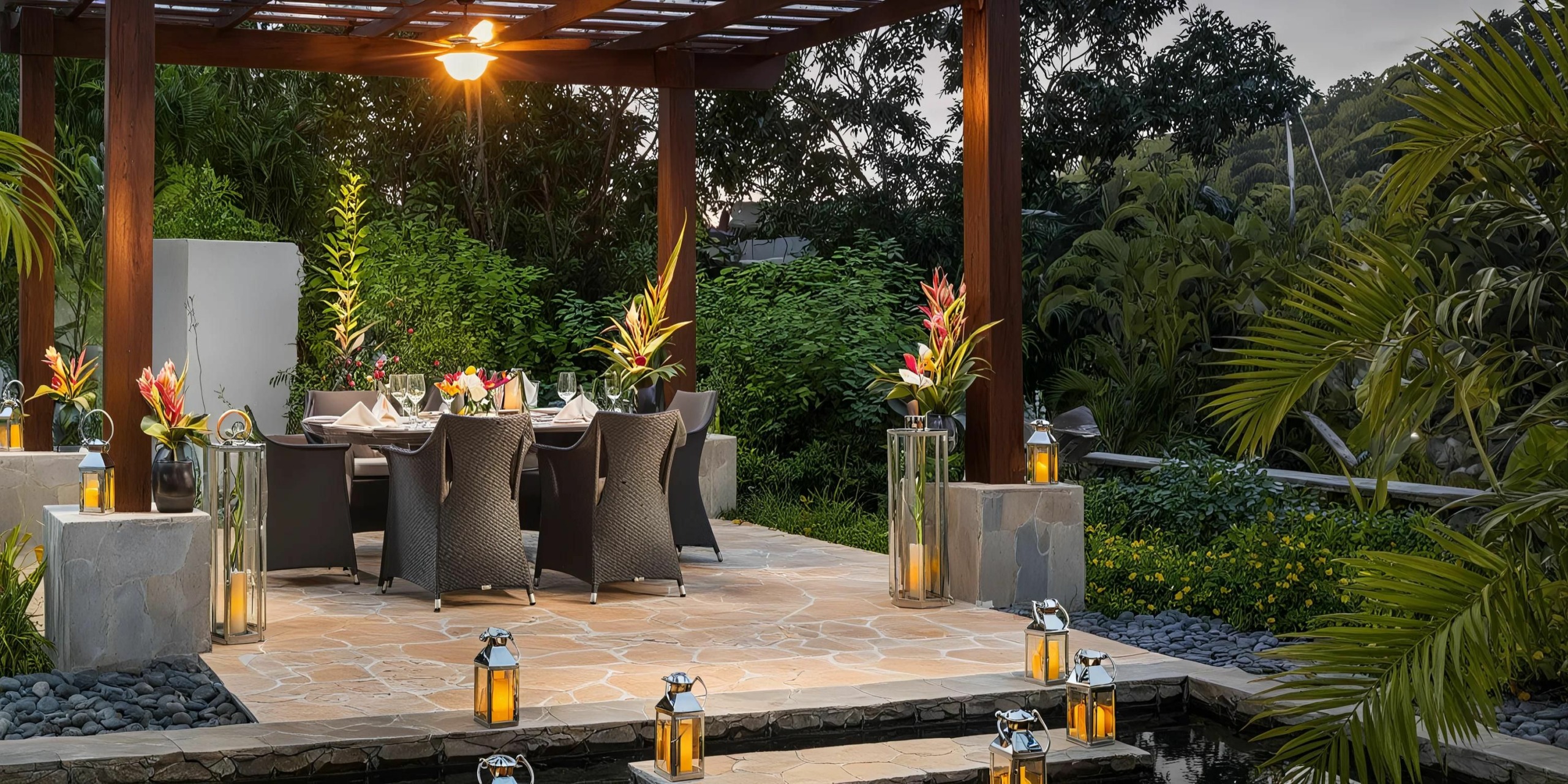 A relaxing dining area at Zoetry Marigot Bay St. Lucia features a wooden pergola amidst lush green surroundings. The table is elegantly adorned with flowers and tableware, while lanterns are strategically placed on the ground around the cozy seating area.