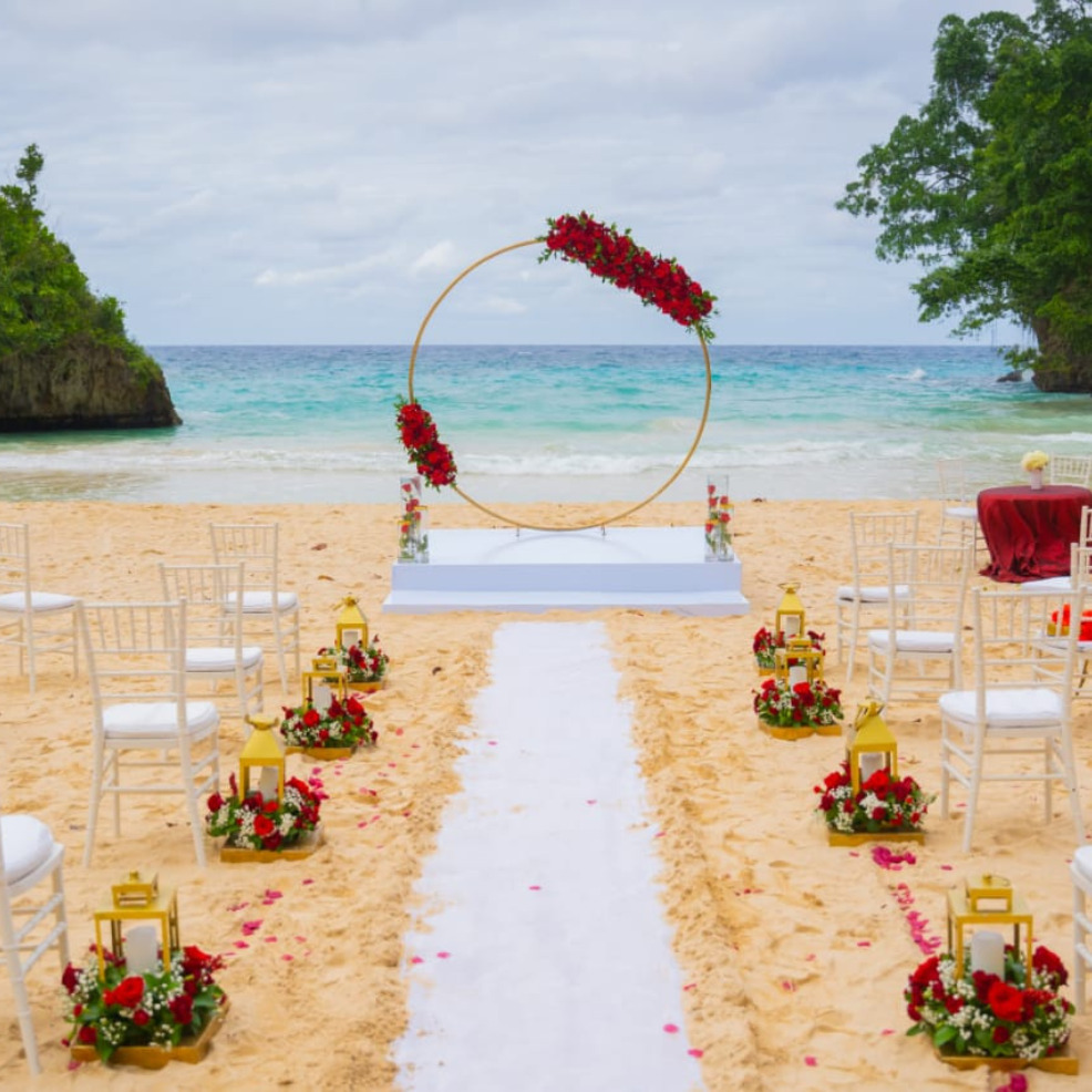 Destination beach wedding setup with a floral arch, white aisle, and chairs on sandy shore facing the ocean.