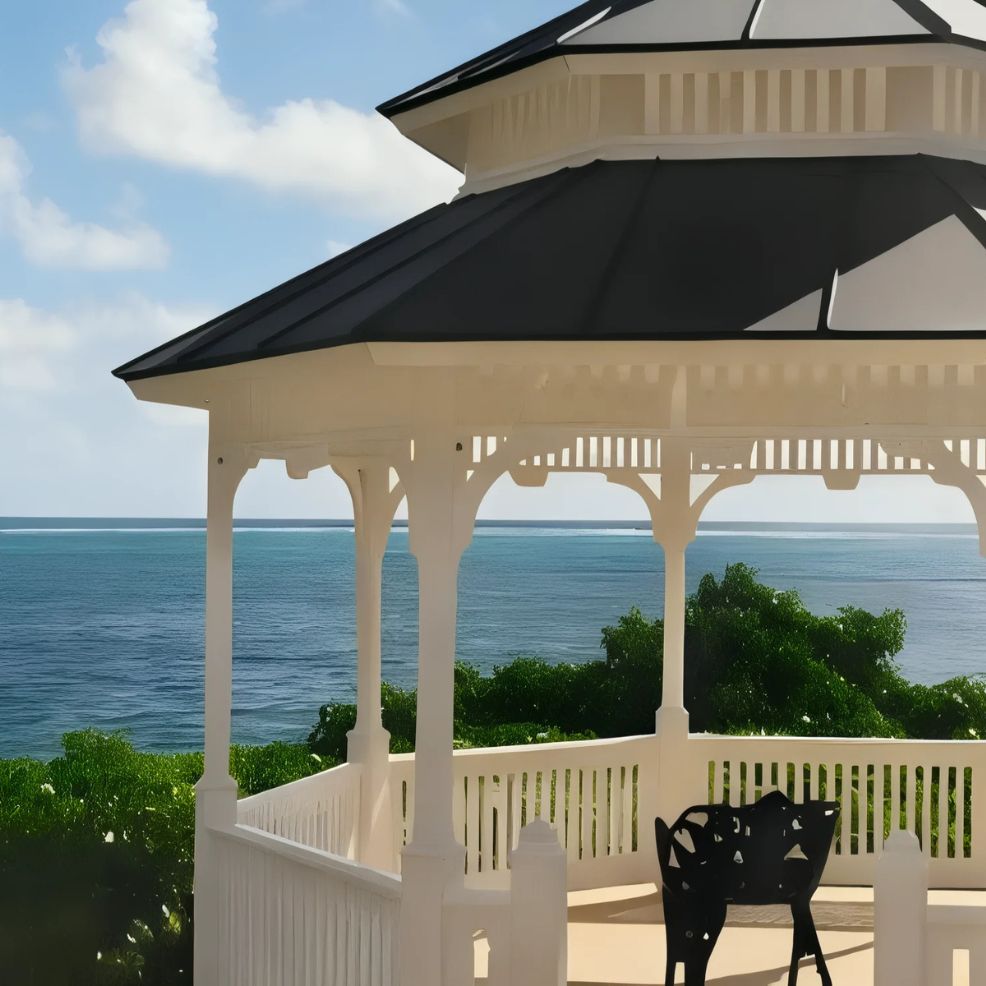 A white gazebo with a black roof overlooks the ocean, surrounded by green foliage under a partly cloudy sky.