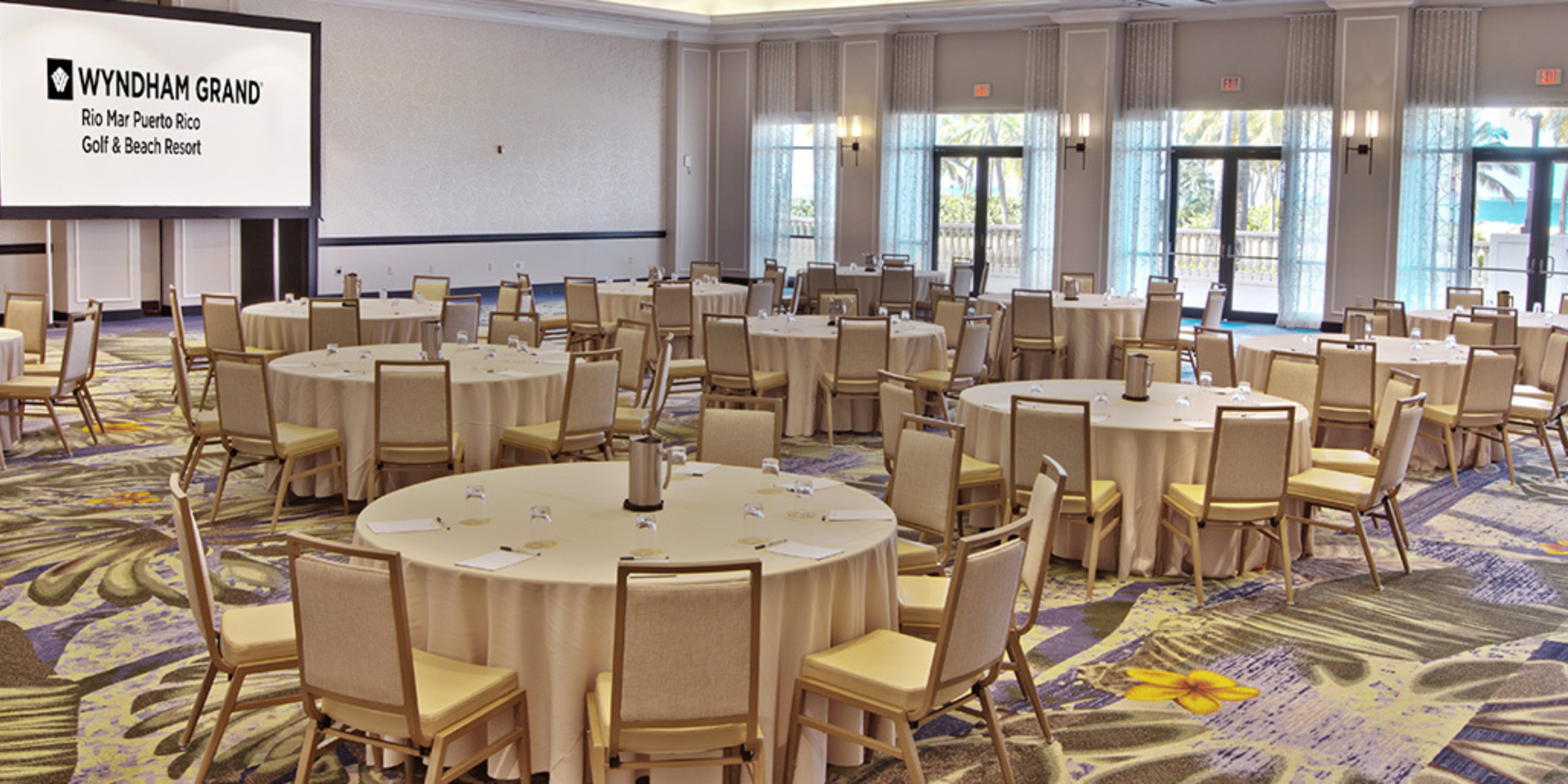 Empty conference room with round tables, beige linens, and chairs, featuring large windows and a screen displaying "destination wedding at Wyndham Grand, Rio Mar Puerto Rico Golf & Beach Resort".