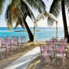 Outdoor beach wedding setup with rows of chairs adorned with pink sashes facing a decorated arch between palm trees, overlooking the blue ocean&mdash;a picturesque scene perfect for Barbados Wedding Packages.