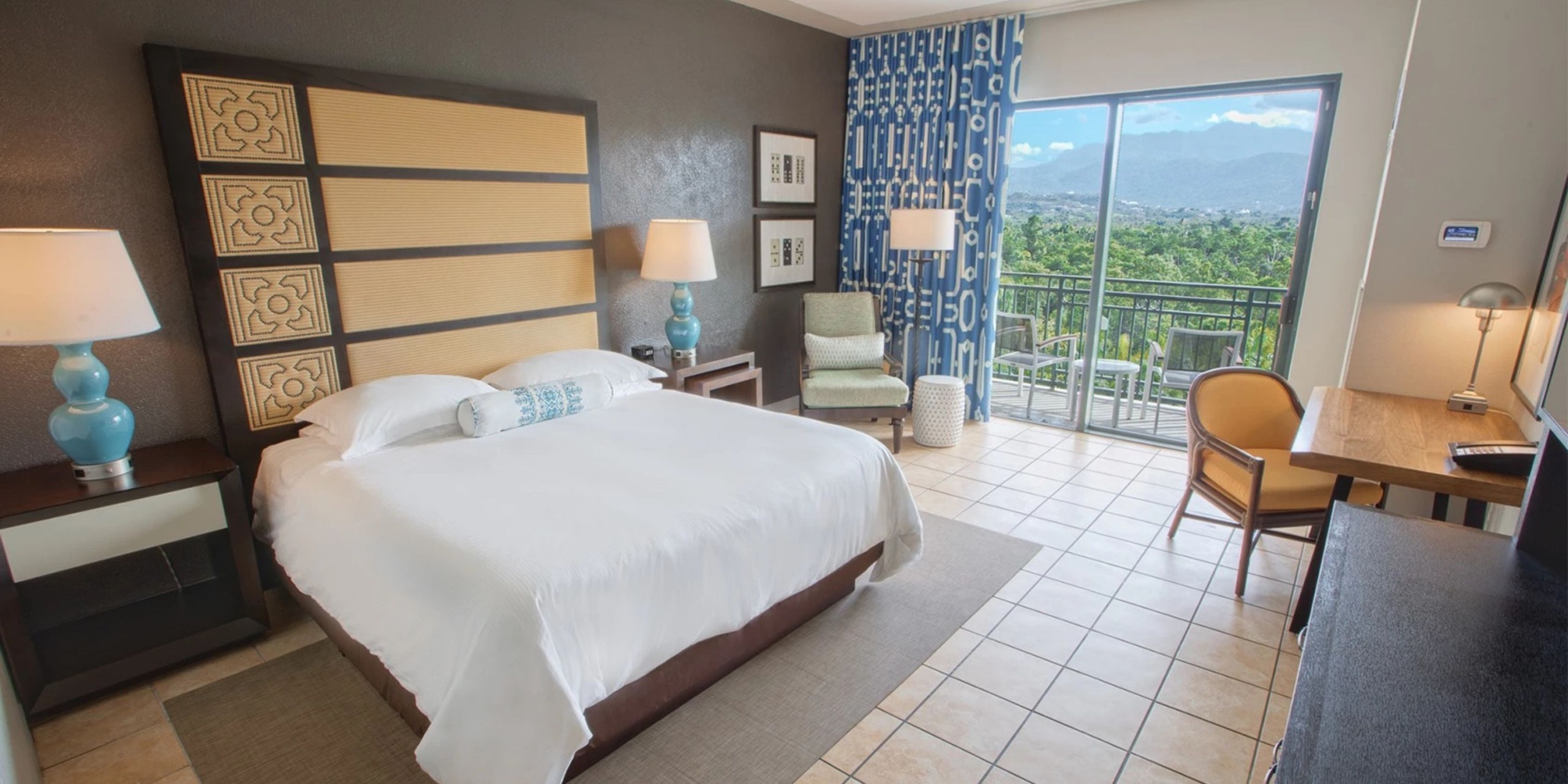 A room at the Wyndham Grand Rio Mar Puerto Rico Golf & Beach Resort features a spacious bed, two lamps, and a desk. There's also a chair positioned by the window that presents views of lush greenery and mountains.