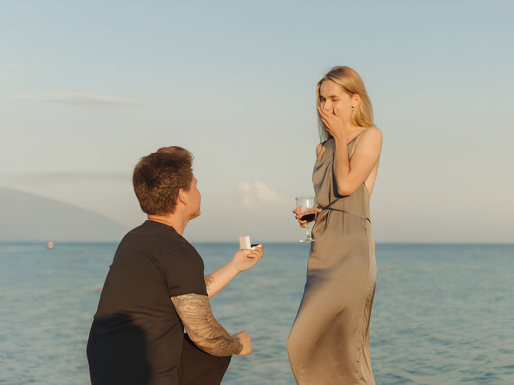 A man kneels and proposes to a surprised woman holding a wine glass—one of the most unforgettable wedding proposal ideas, perfect for announcing your plans for a dream destination wedding.