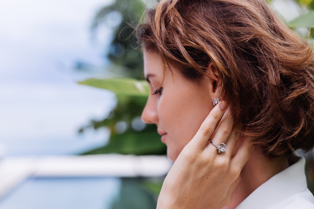 A woman adjusts her earring, highlighting a stunning lab-grown diamond ring—an elegant choice for a destination wedding—amid lush greenery.