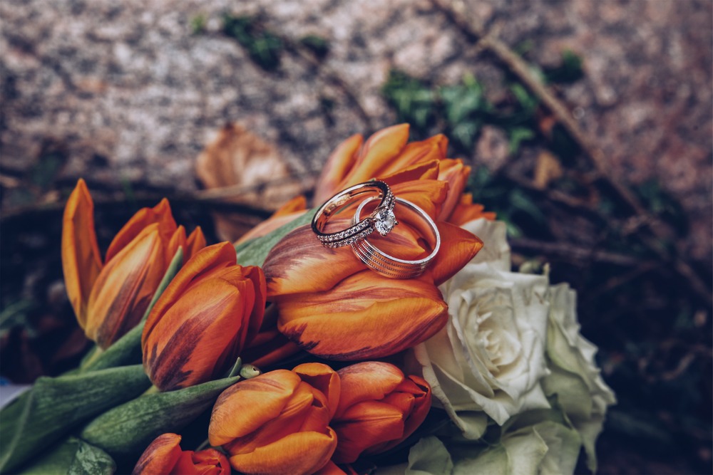 Two silver wedding bands and a lab-grown diamond ring are elegantly displayed atop vibrant tulips and a white rose, set against a stone backdrop—perfect inspiration for your destination wedding.