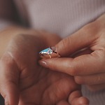 A person delicately holds a gold lab-grown diamond ring between their thumb and finger, perhaps envisioning it as the perfect choice for a wedding or even a dream destination wedding.