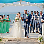 Bride and groom walking down the aisle by the beach, smiling, with guests looking on, some clapping, in a sunny destination wedding.
