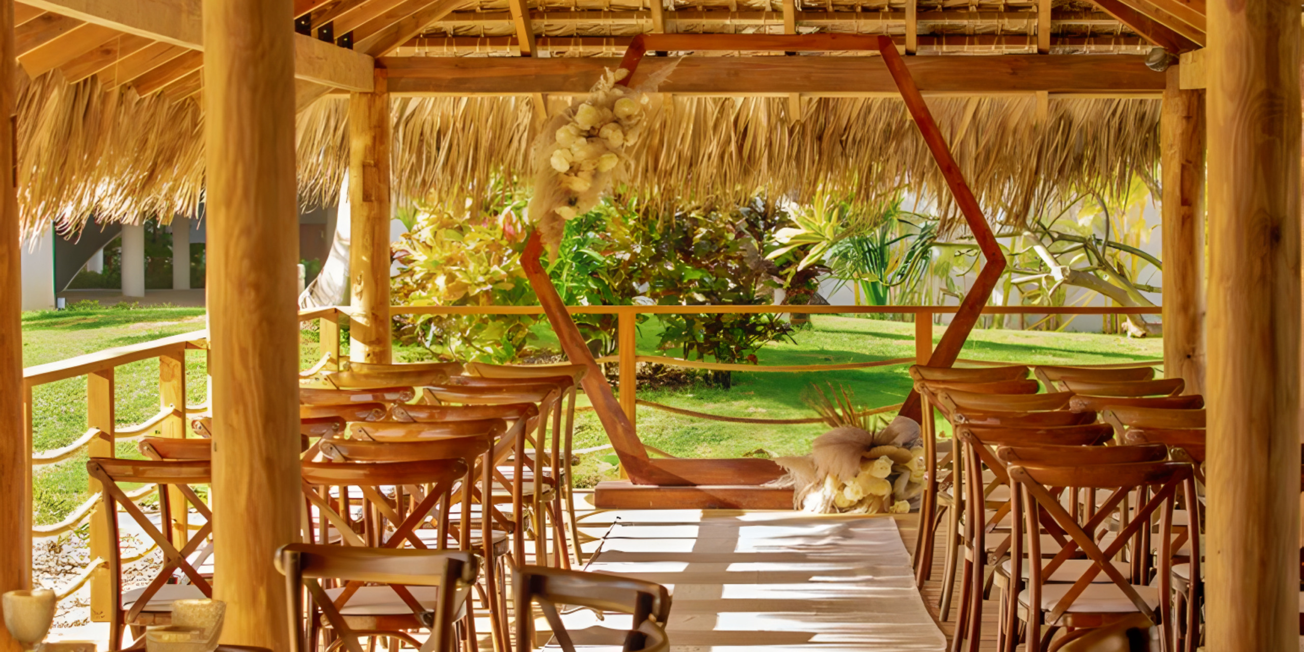 A cozy, open-air wooden bar with thatched roof, overlooking a lush garden—perfect for a destination wedding. Wooden stools line the bar under hanging dried flowers.