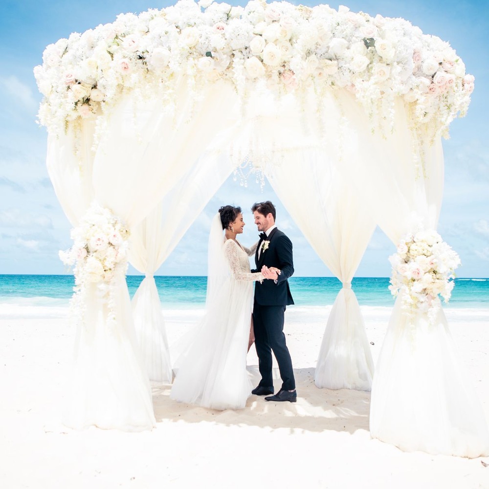 A bride and groom exchange vows beneath a floral arch by the ocean—a picture-perfect scene that captures the essence of a classic destination wedding.