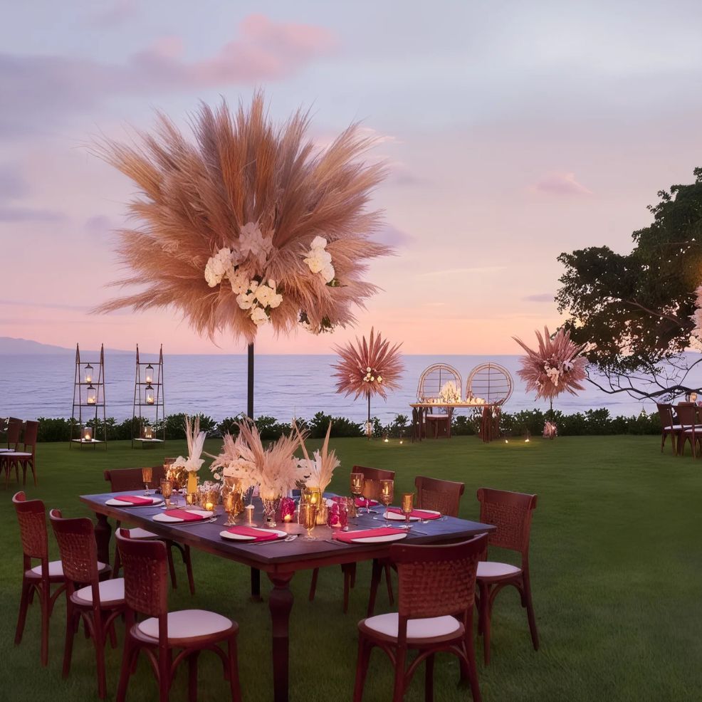 Outdoor dining setup by the sea at sunset, featuring tables with floral centerpieces and wicker chairs on a green lawn.