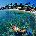 A woman snorkeling in clear blue water near a sandy shore with tropical huts and palm trees, enjoying a destination wedding.