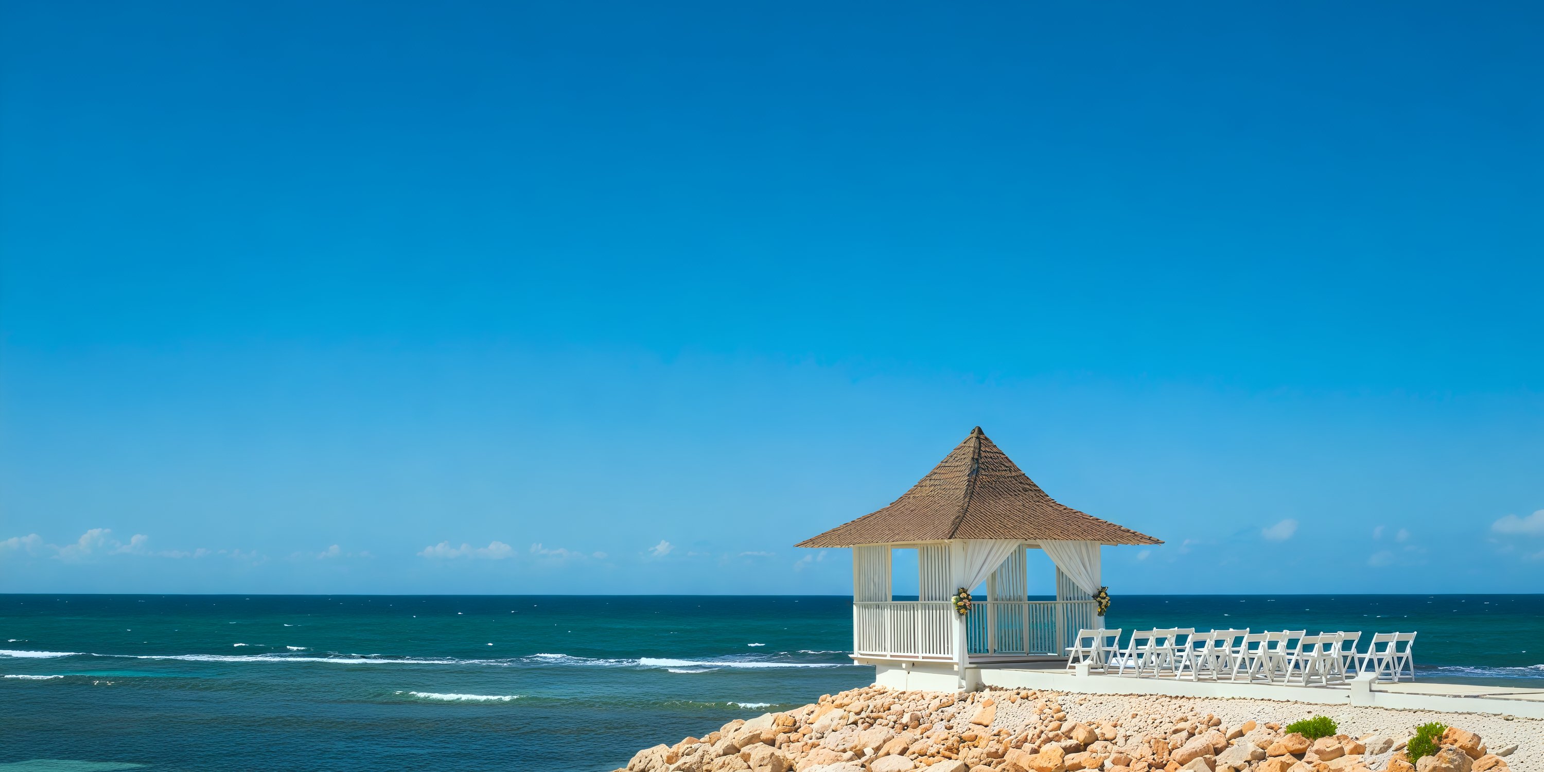 Ocean Coral Spring with its picturesque white gazebo and chairs set on a rocky shore creates a perfect backdrop for a destination wedding under the clear blue sky.