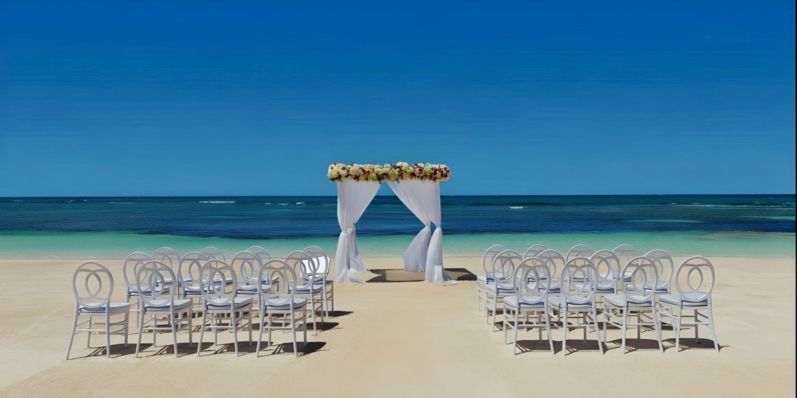 Destination wedding setup with a white floral arch and rows of white chairs on sandy shore facing the ocean.