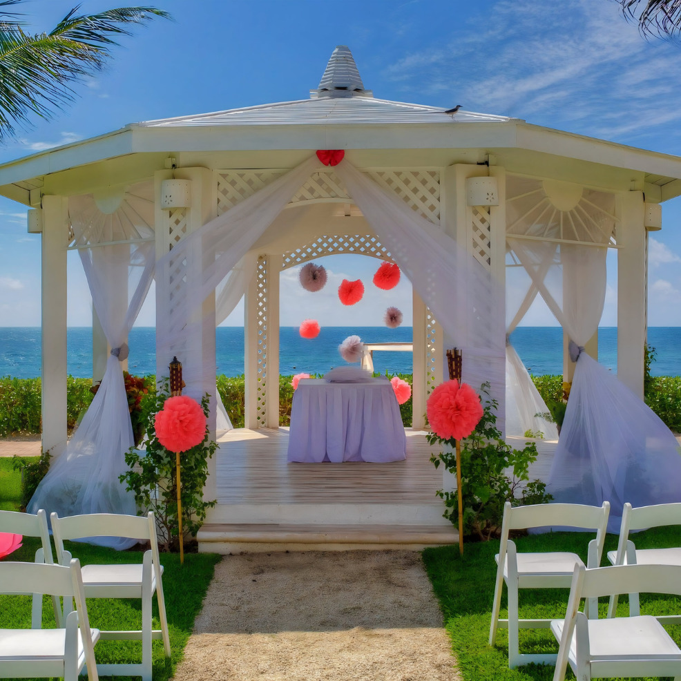 A destination wedding gazebo adorned with white drapes and pink flowers, flanked by white chairs, overlooking the ocean.