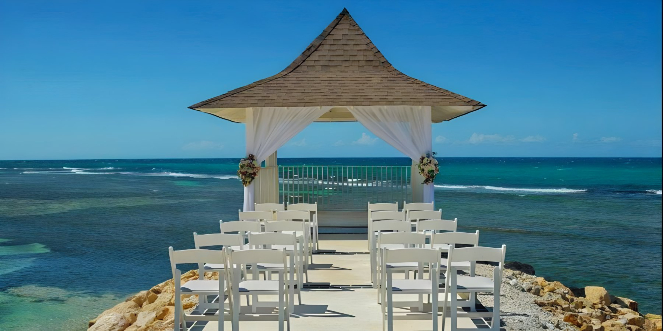 White chairs arranged for a destination wedding ceremony under a wooden gazebo, with a clear blue sky and ocean in the background.