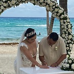 A bride and groom sign a document at their destination wedding beneath a floral arch by the sea.