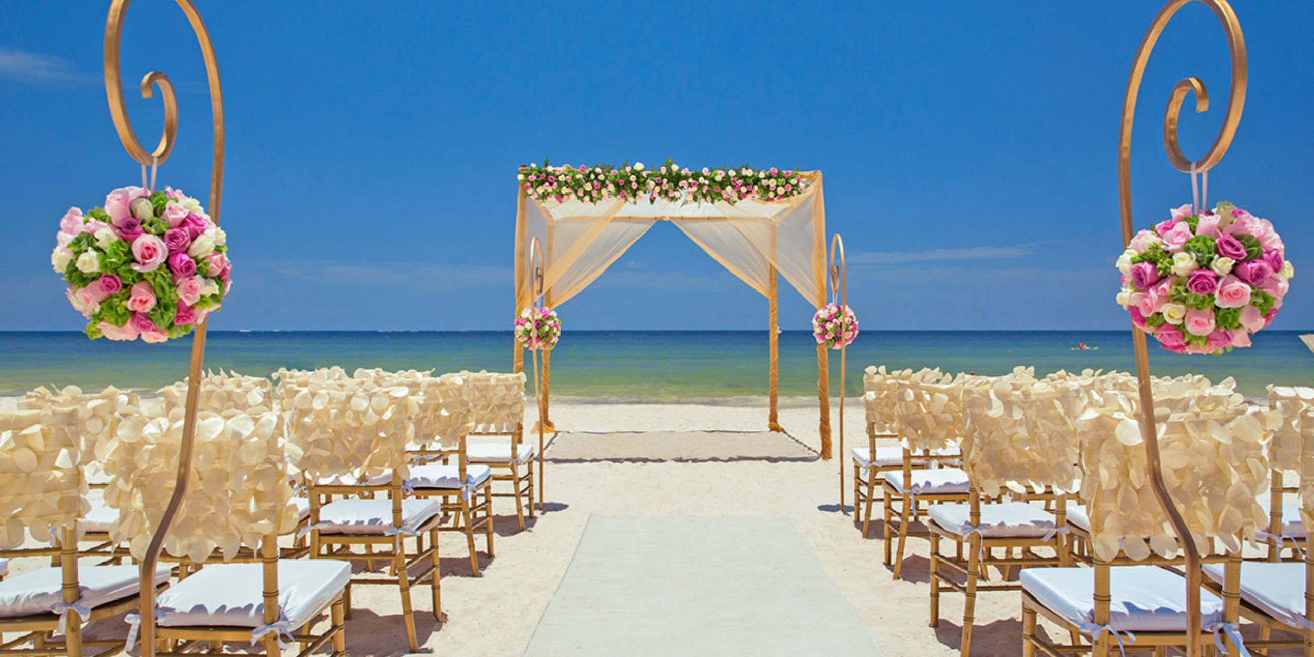 Destination wedding setup with a floral arch and rows of white chairs under a clear blue sky.