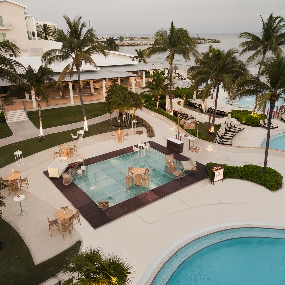 Poolside area with palm trees, tables, and a glass dance floor by the ocean set for a destination wedding.