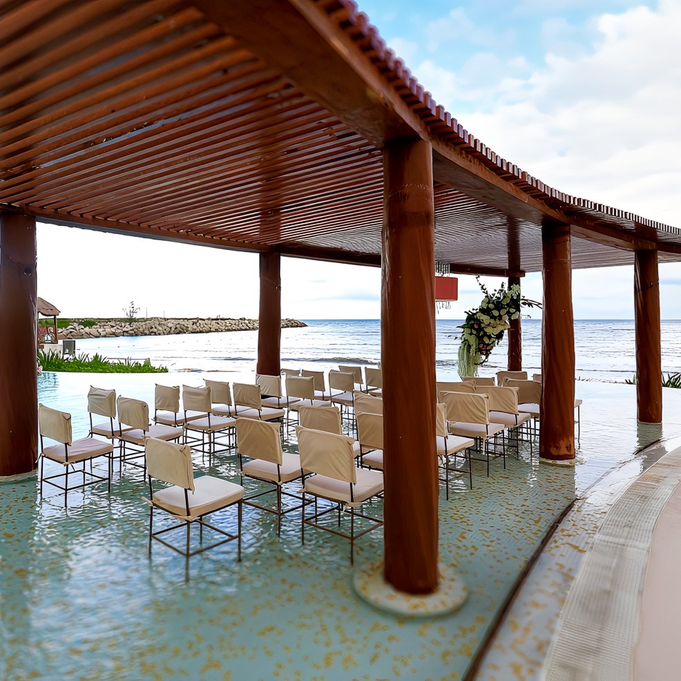 Rows of chairs arranged under a wooden pergola by the sea, ready for a destination wedding ceremony.