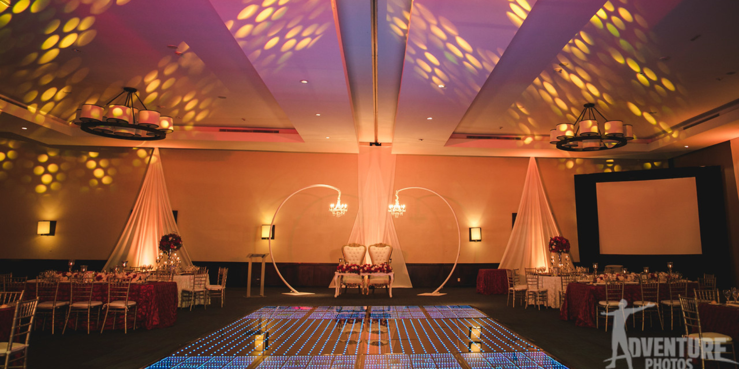 Elegant destination wedding venue set for a reception with a lit dance floor, patterned lights on the ceiling, and draped chairs under arches.