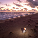Aerial view of a person walking alone on a sandy beach at sunset, with waves approaching the shore and a cloudy sky above, capturing the tranquil atmosphere before a destination wedding.