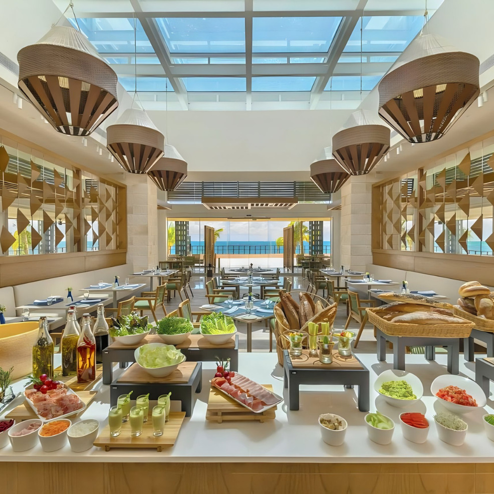 Bright, spacious buffet restaurant ideal for destination weddings, with large windows, modern decor, and an array of fresh food displayed on tables under hanging wicker lamps.