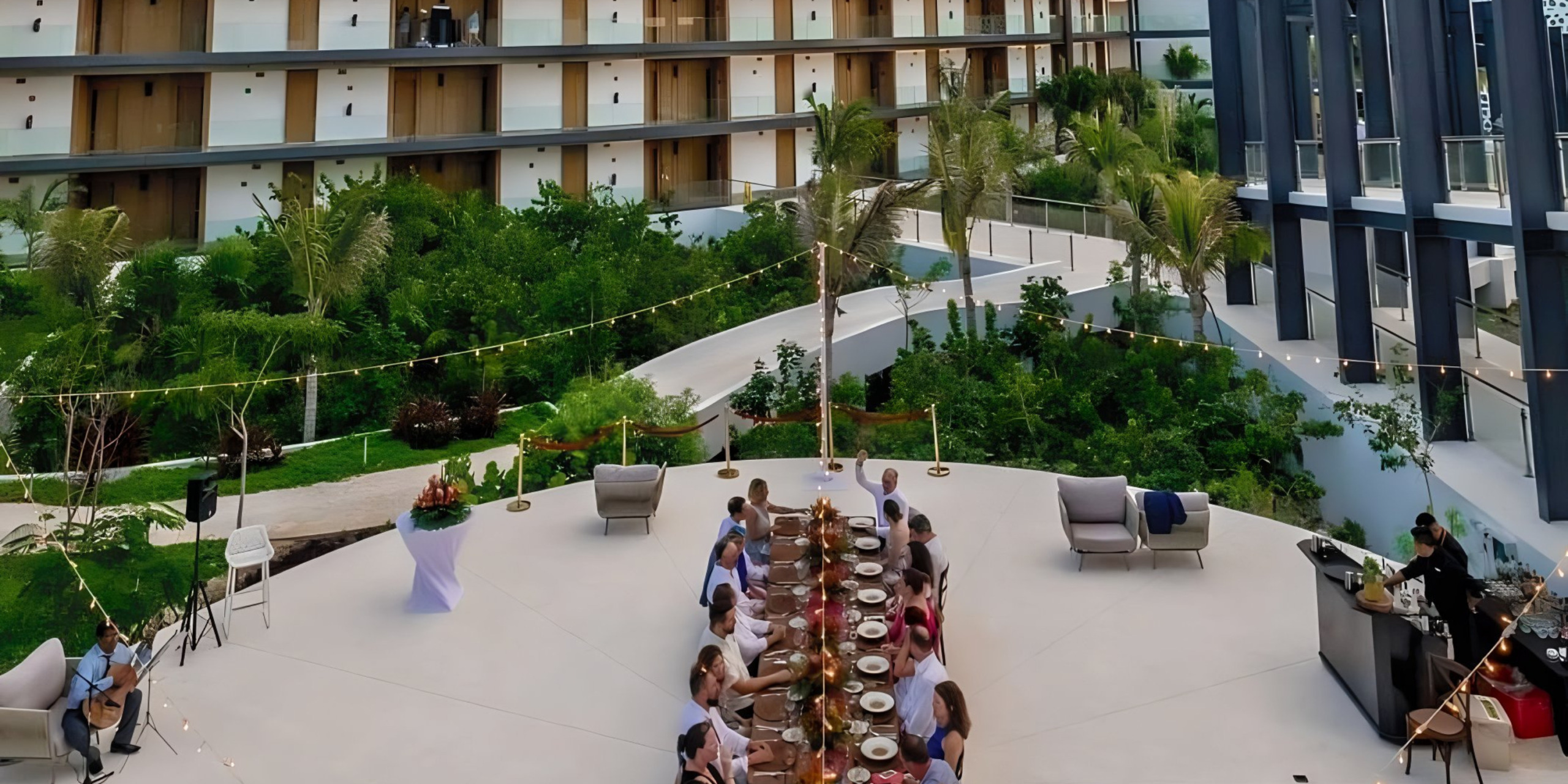 A group of people dining outdoors at a circular table in a garden terrace surrounded by modern residential buildings, celebrating a destination wedding.