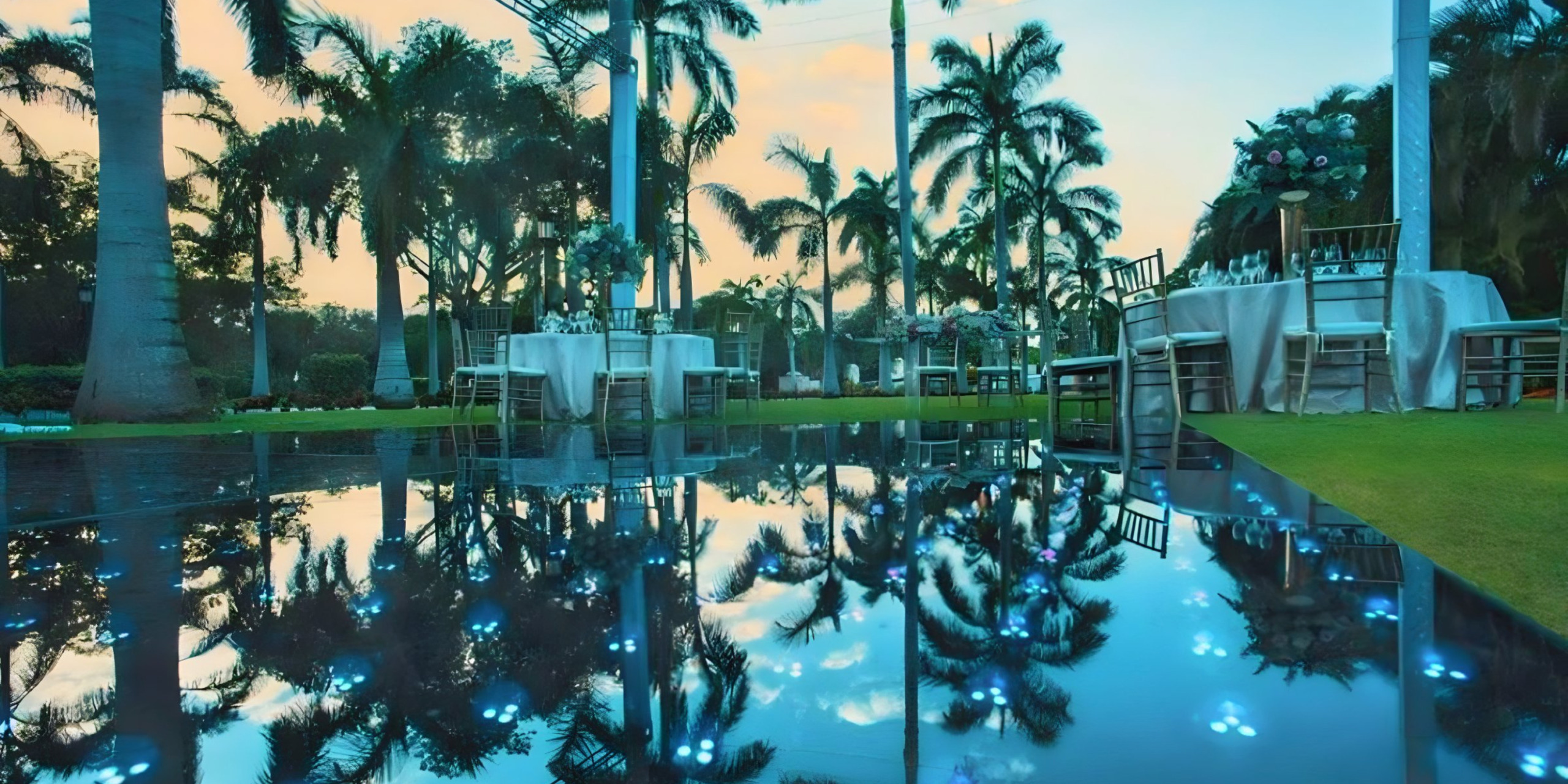 Destination wedding venue at twilight with tables set for a reception, palm trees, and a reflective wet floor enhancing the serene ambiance.