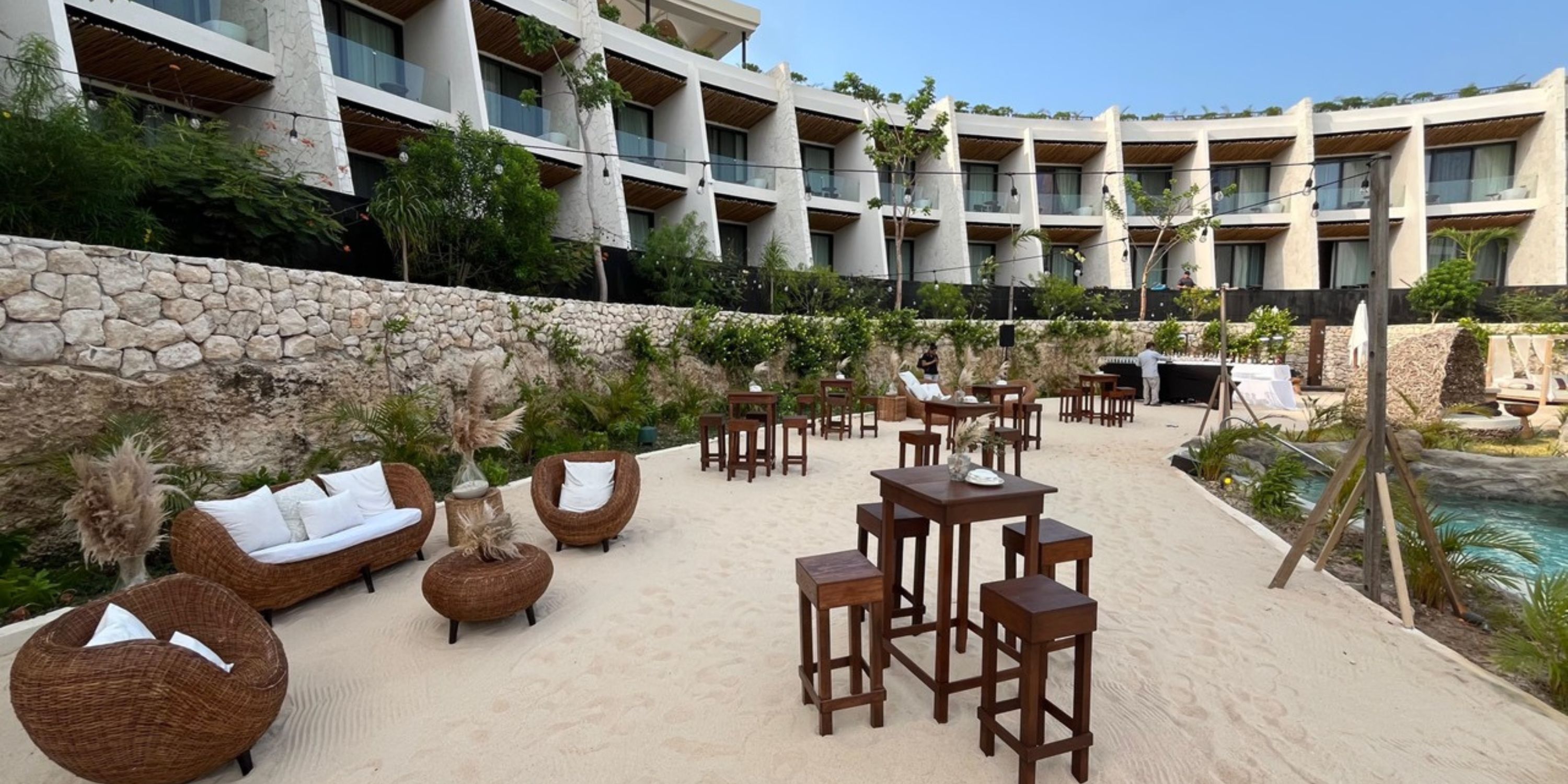 Wicker chairs and tall tables on sand outside the modern, curved Secrets Tulum hotel for a wedding.