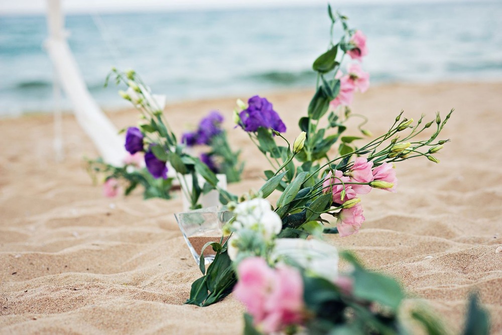 Elegant flower arrangements in glass vases adorn the sandy beach, with the sparkling ocean providing a stunning backdrop—perfect for a romantic destination wedding.