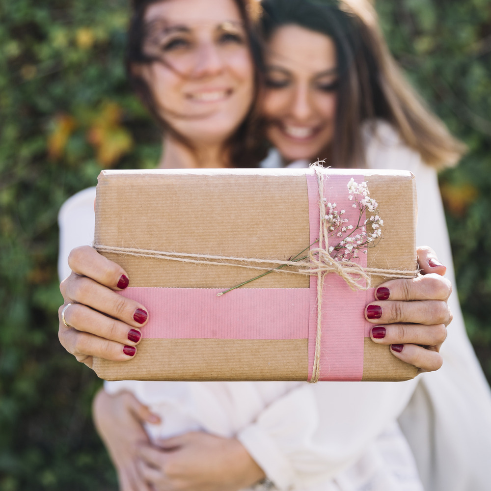 Two women share a warm embrace at a destination wedding as one hands the other a beautifully wrapped wedding gift adorned with brown paper and finished with a pink ribbon.