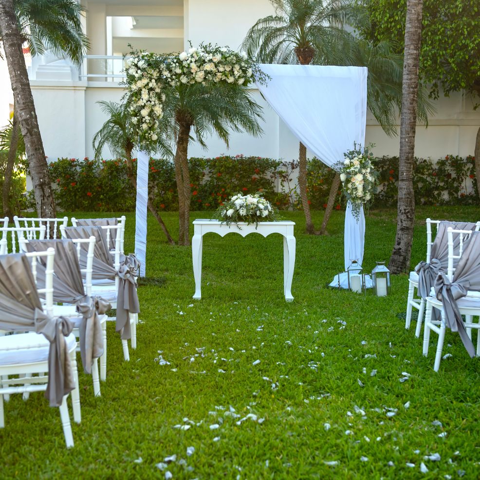 Destination wedding ceremony arranged outdoors with white chairs, floral arch, and table on lush lawn.